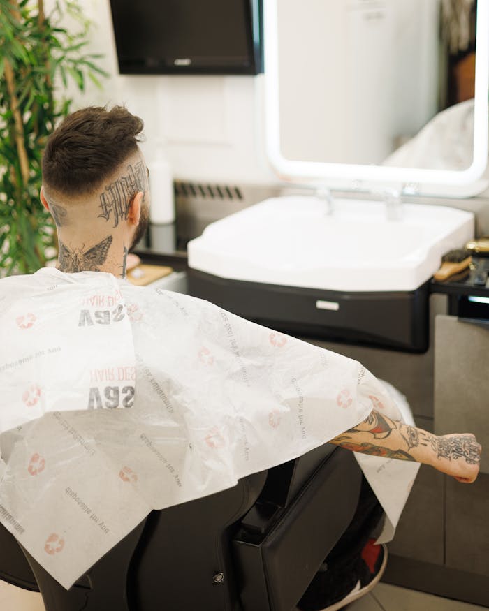 The Art of Drawing Readers In: Your attractive post title goes here A tattooed man sits in a barber shop chair wearing a cape, with modern decor and a mirror in the background.